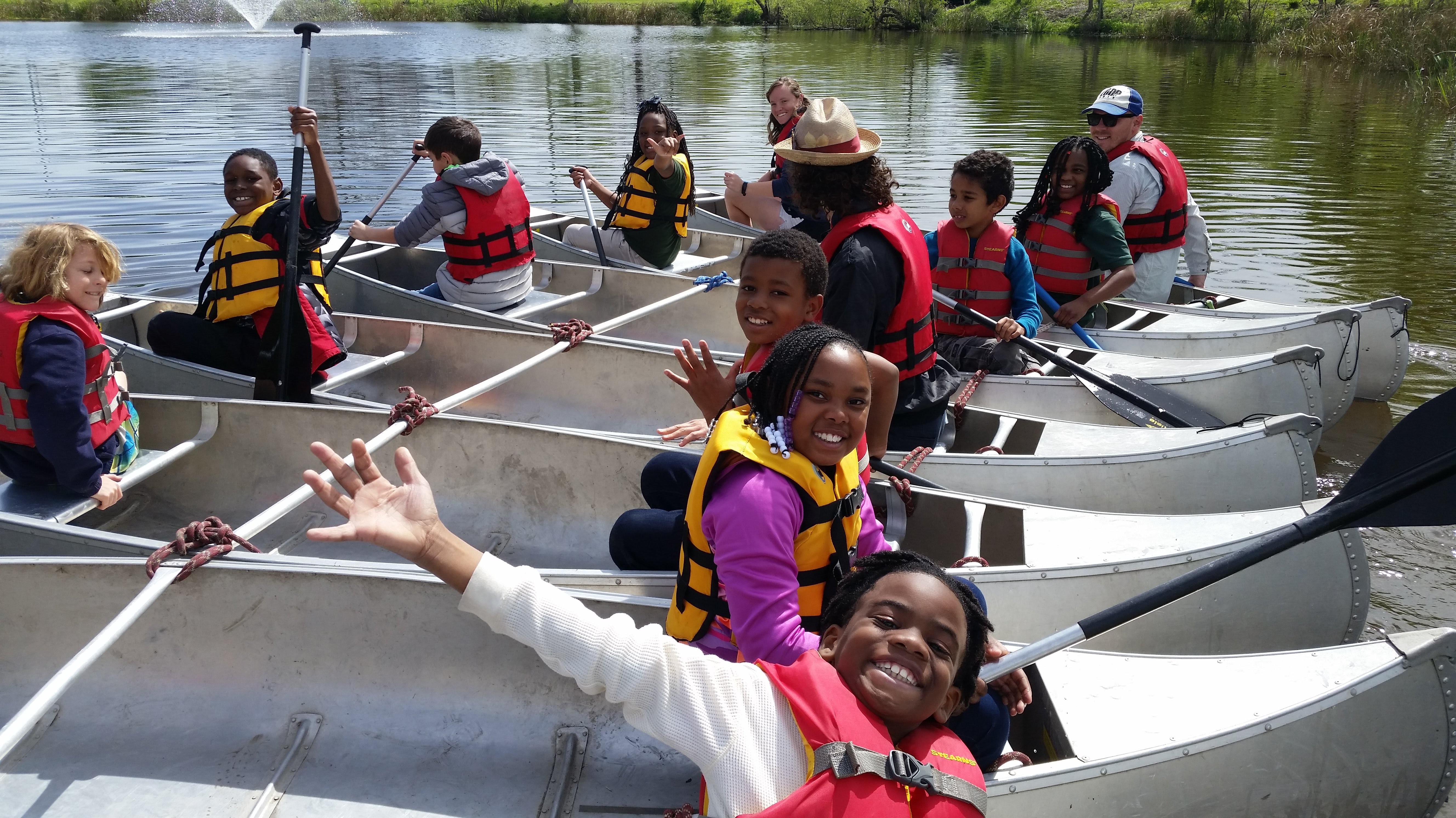 A group of kids wave and smile at the camera from canoes